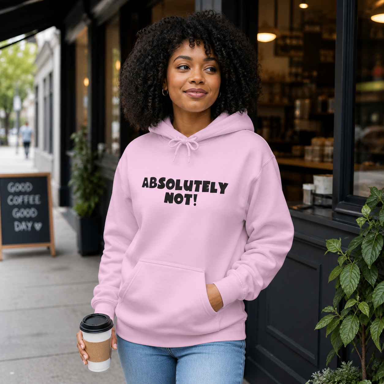 Woman wearing a light pink Absolutely Not graphic hoodie outside a neighborhood cafe