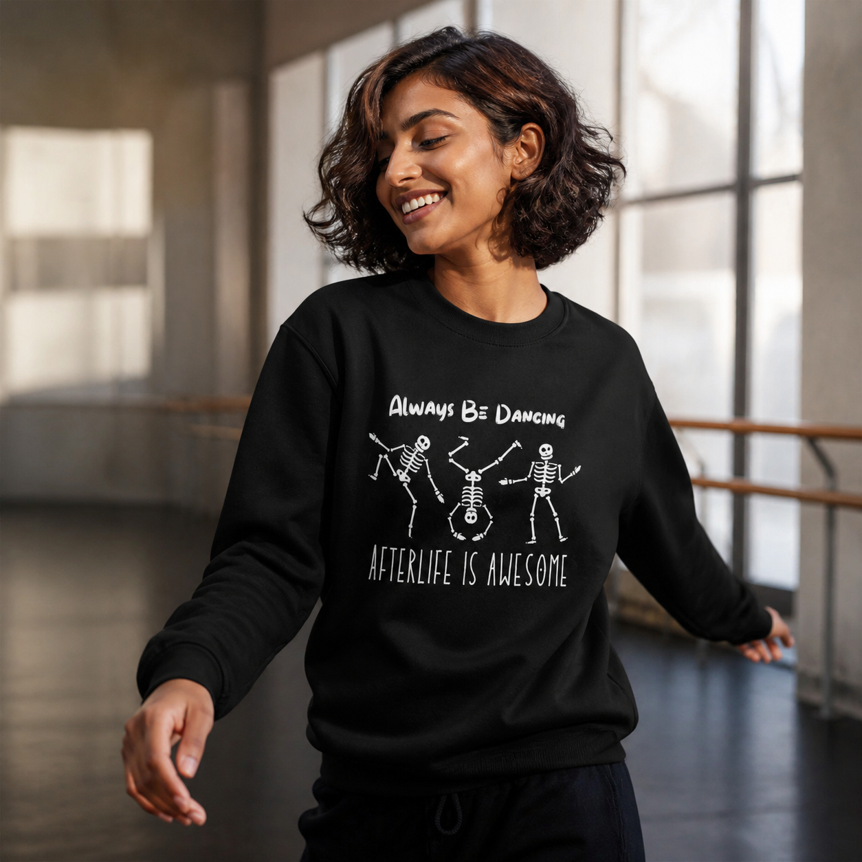 Young woman wearing black Always Be Dancing Afterlife Is Awesome skeleton sweatshirt in a dance studio