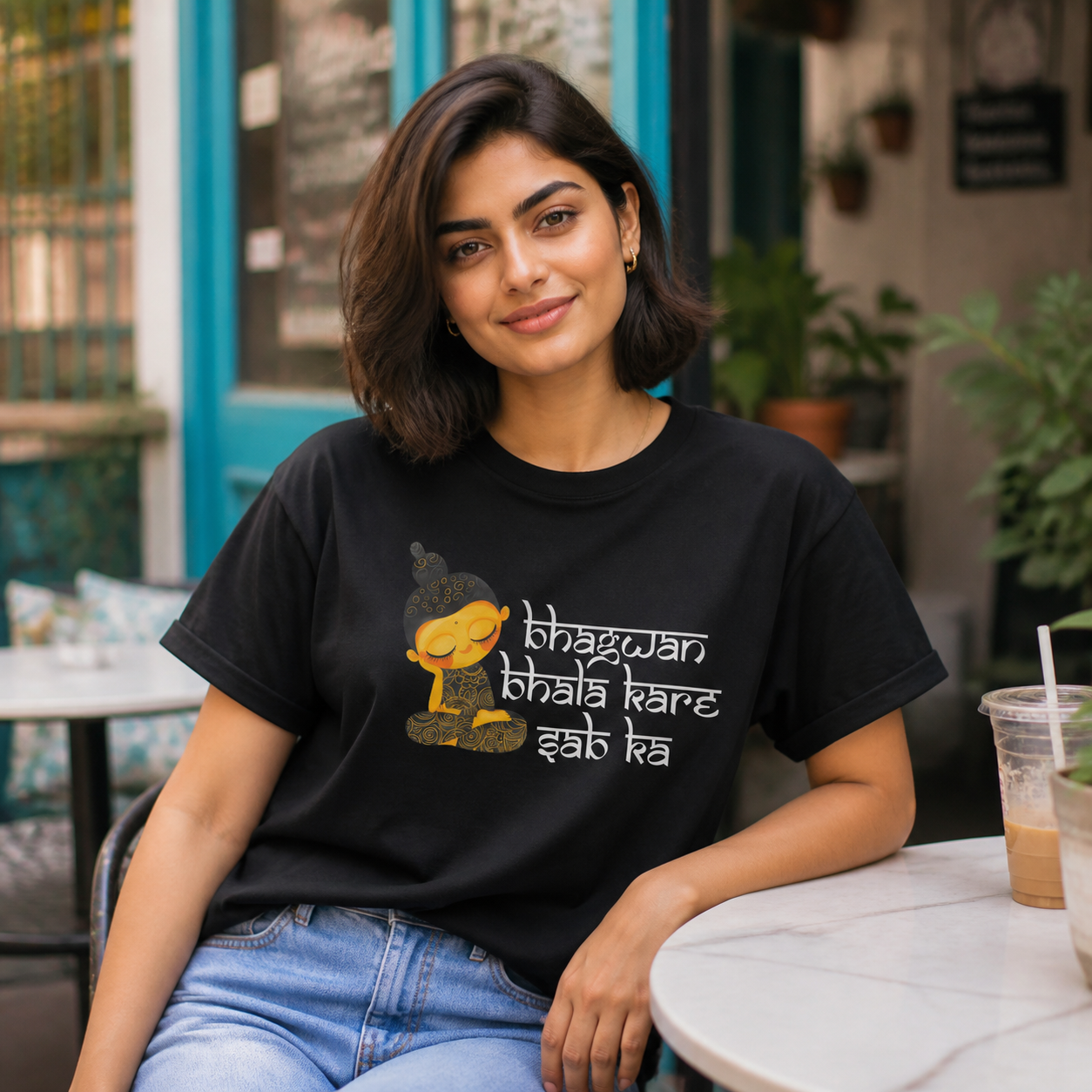 Young North Indian woman wearing Bhagwan Bhala Kare Sab Ka graphic T-shirt at an urban cafe
