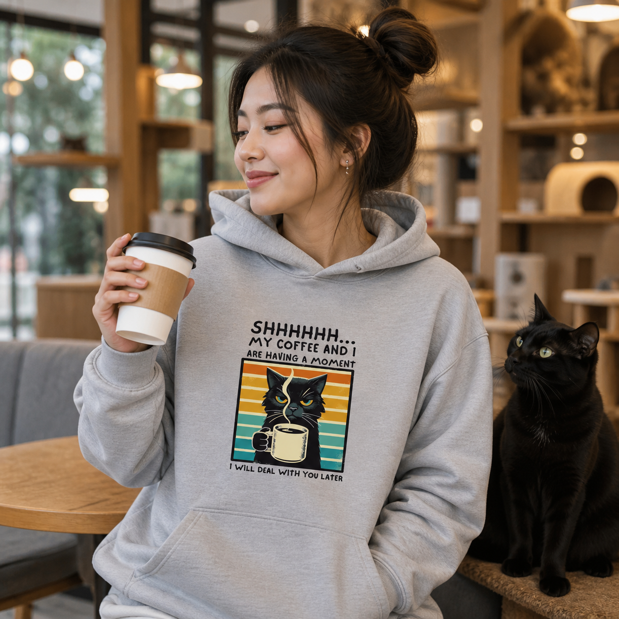 Woman wearing gray Coffee Cat hoodie with a black cat in a modern café