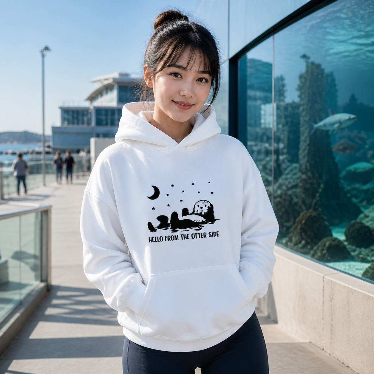 Young woman wearing Hello From the Otter Side graphic hoodie at an aquarium promenade