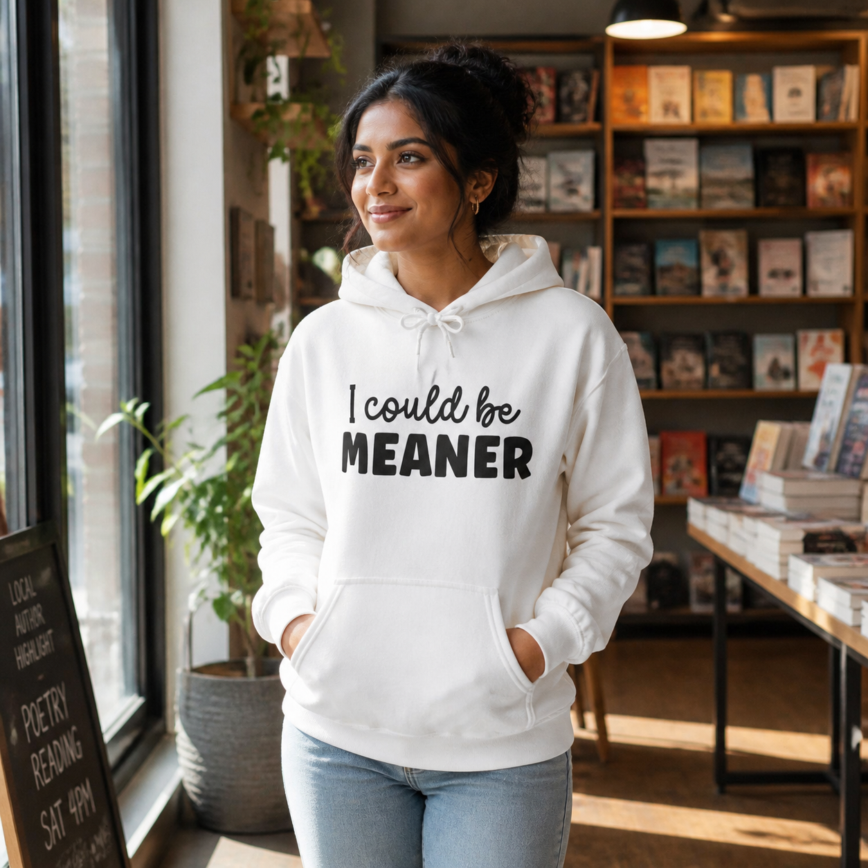 Young South Asian woman wearing a white I Could Be Meaner hoodie in a bookstore cafe
