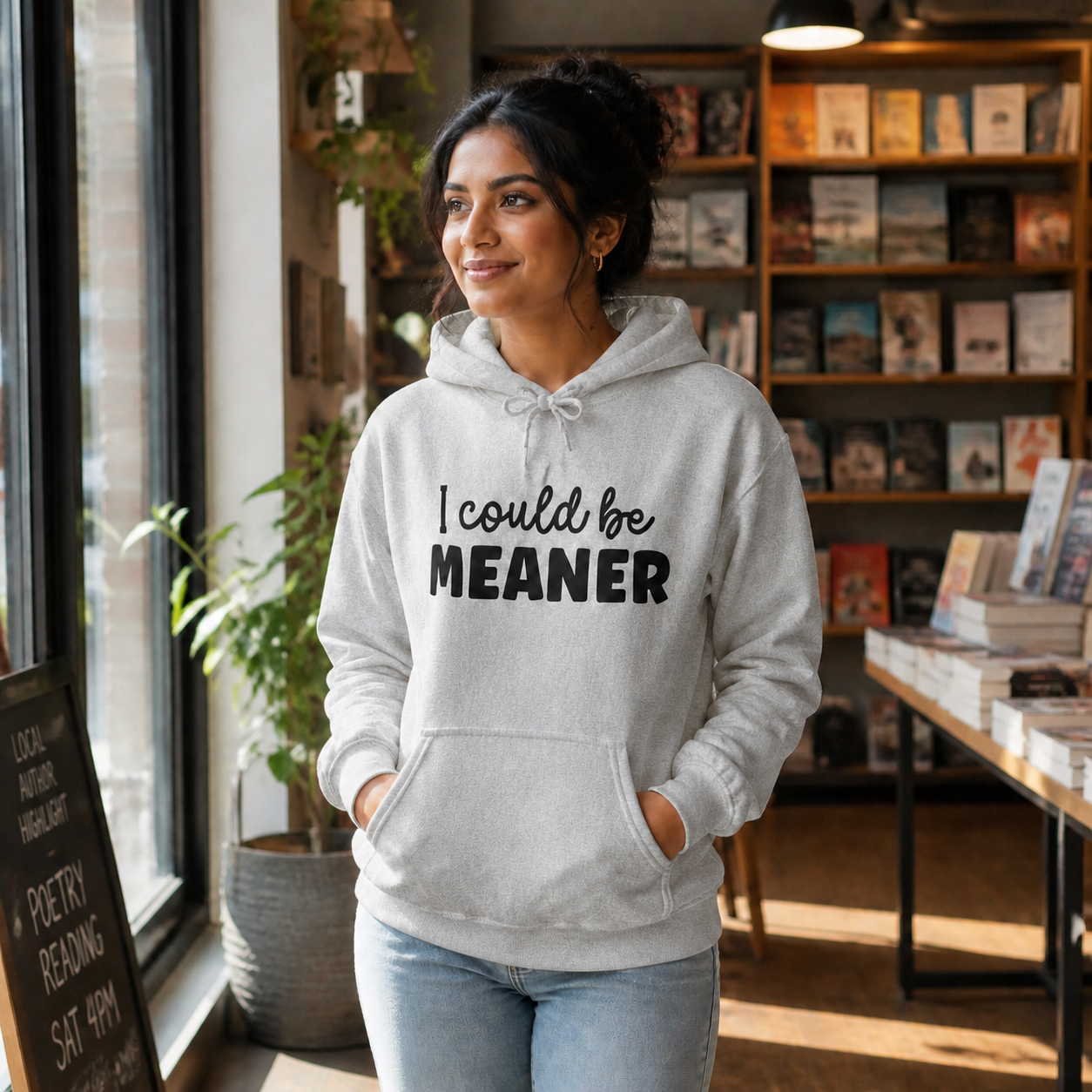 Young South Asian woman wearing a Sport Grey I Could Be Meaner hoodie in a bookstore cafe