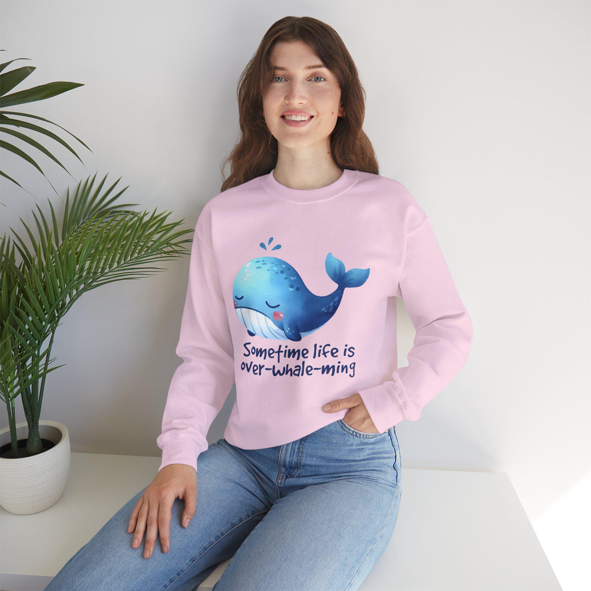 Woman wearing pink 'Sometime Life Is Over-Whale-ming' sweatshirt with whale design, sitting beside a plant.