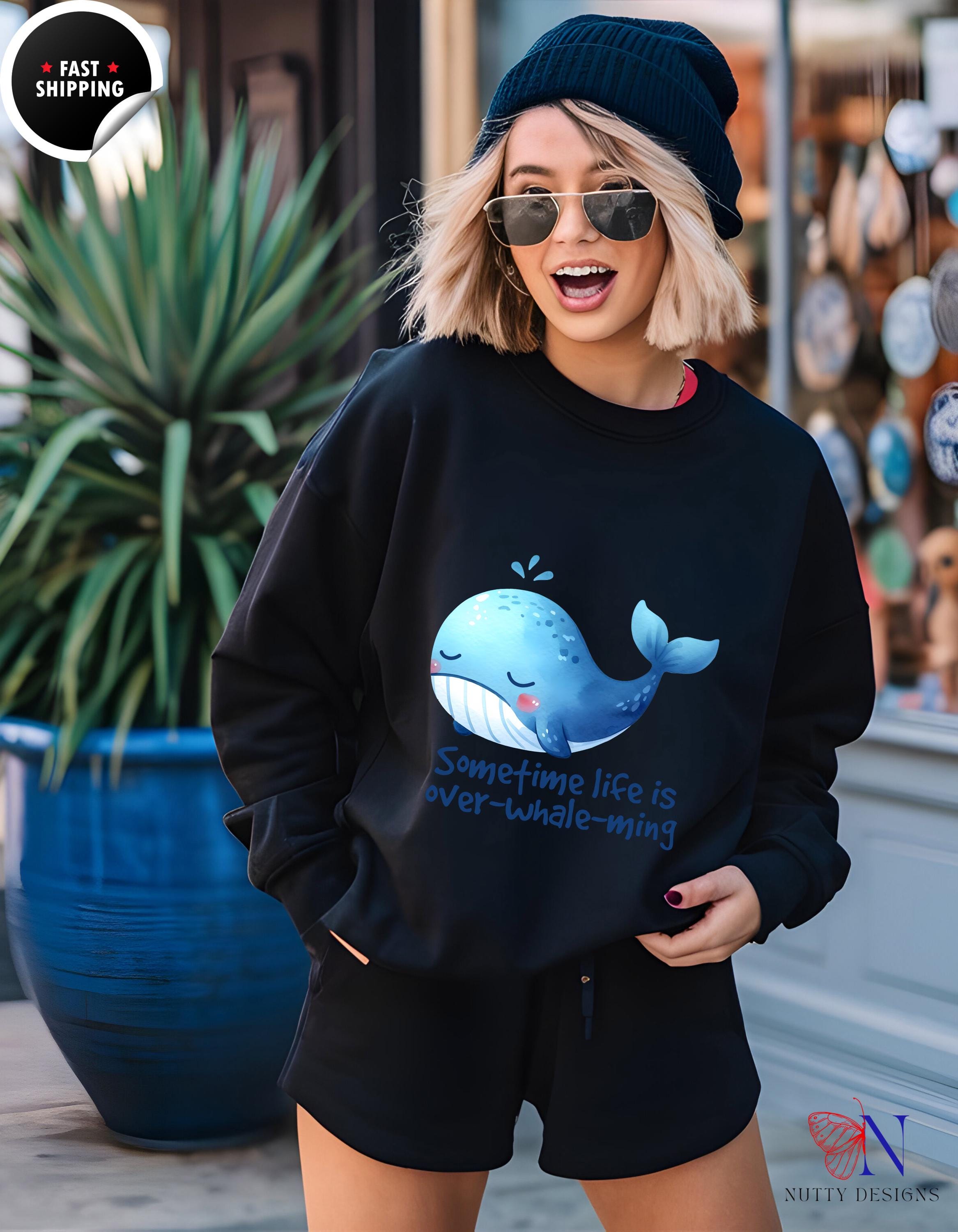 Smiling woman wearing 'Sometimes life is Over-Whale-ming' sweatshirt outdoors, showcasing positive graphic design.