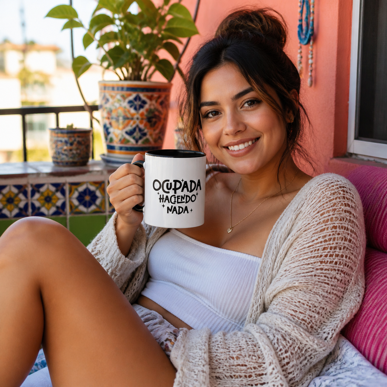 Young Latina woman holding black accent Ocupada Haciendo Nada coffee mug on a colorful balcony
