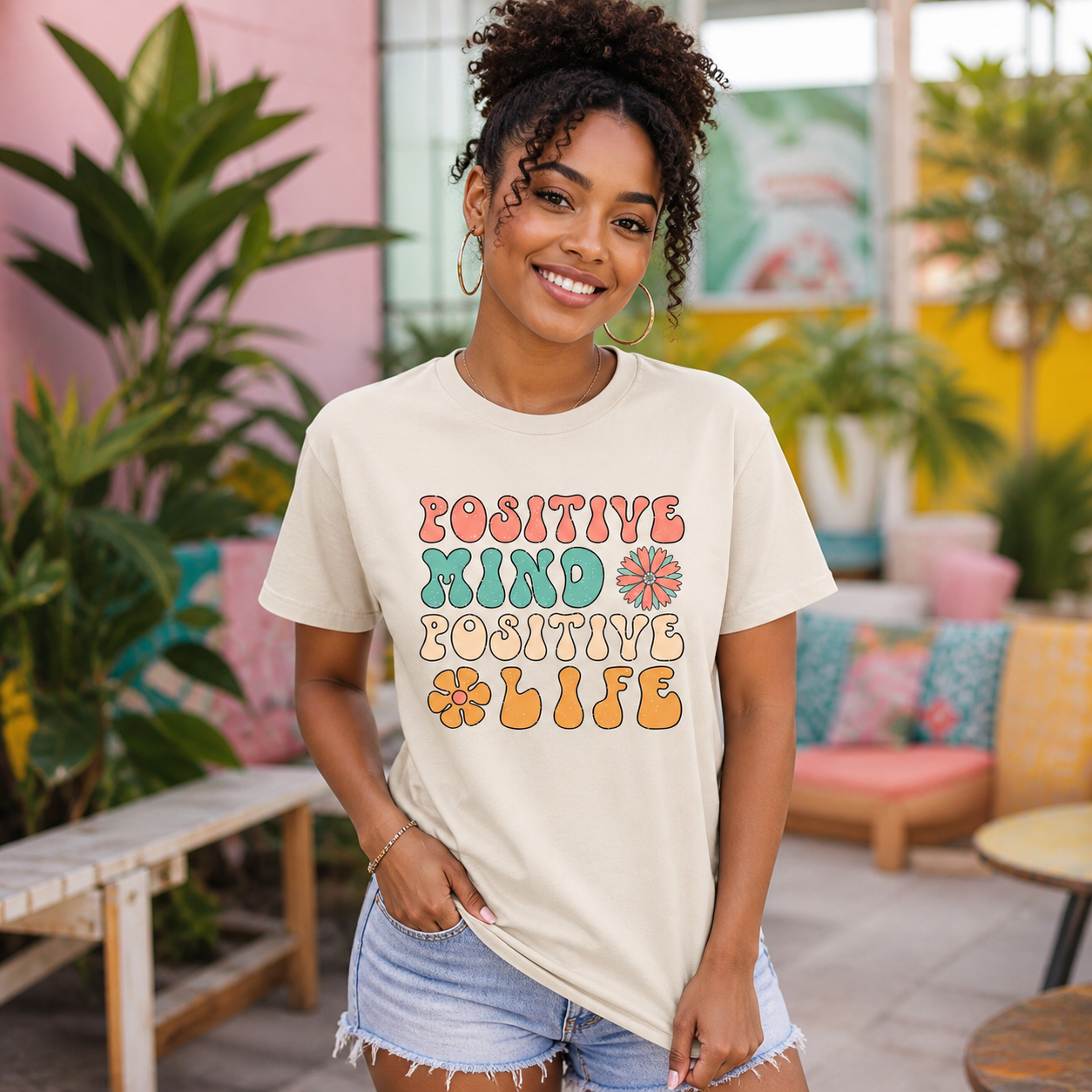 Young woman wearing Positive Mind Positive Life retro graphic T-shirt in a colorful cafe courtyard