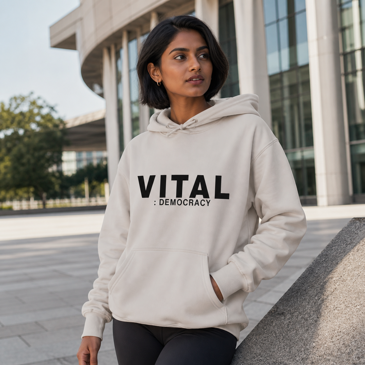 Young woman wearing stone Vital Democracy graphic hoodie at a modern civic plaza