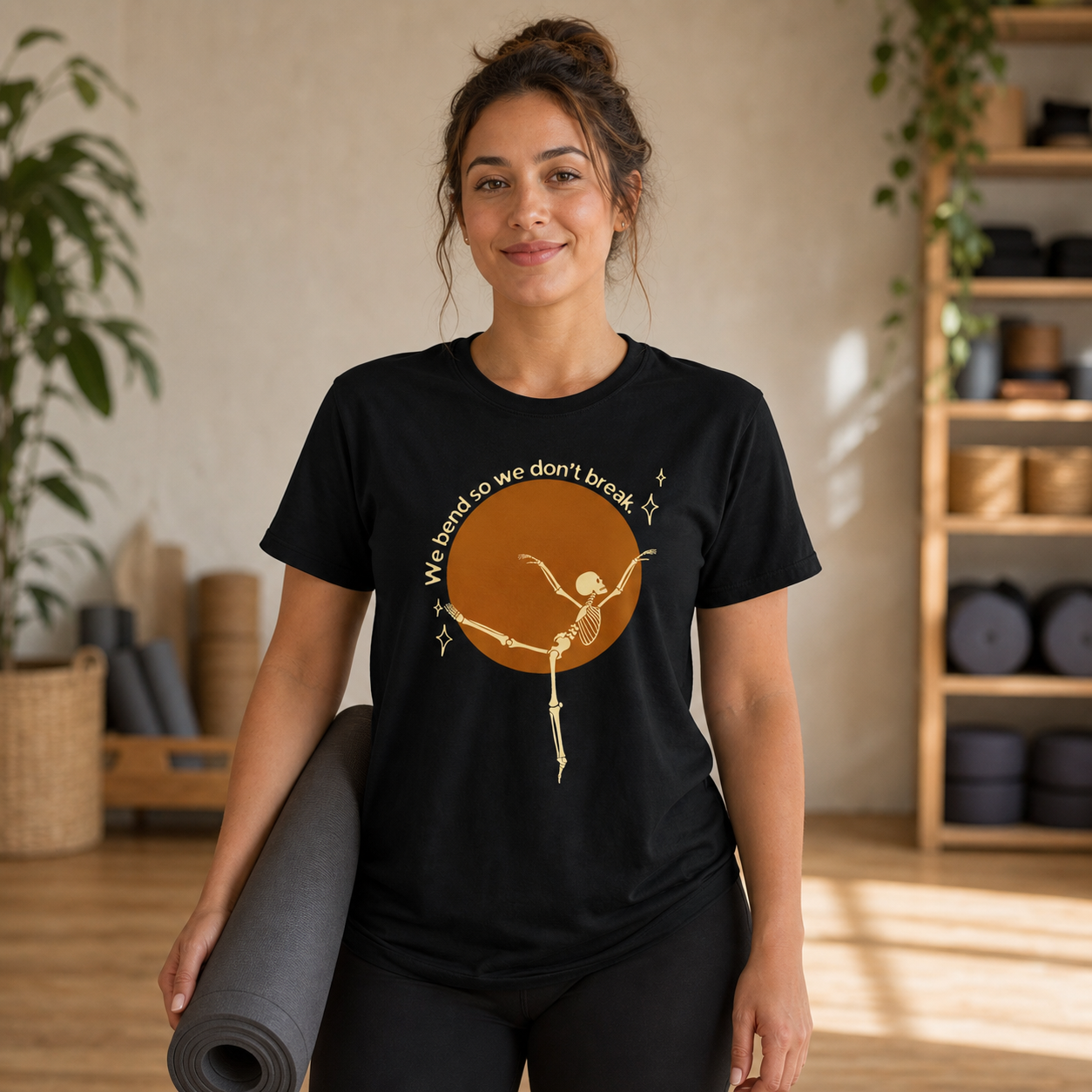 Woman holding a yoga mat wearing a black We Bend So We Don't Break skeleton yoga T-shirt
